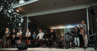 A group of performers on stage at the Nebraska Crossroads Music Festival, playing instruments and singing for the audience.