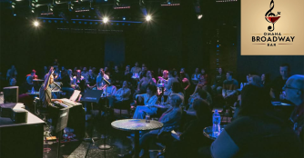 A lively crowd watches a singer at Omaha Broadway Bar, with a piano player in the background under warm stage lighting.