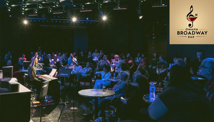 A lively crowd watches a singer at Omaha Broadway Bar, with a piano player in the background under warm stage lighting.