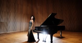 Joyce Yang poses beside a grand piano in an elegant concert setting, marking her performance in the Omaha Symphony’s season finale. She wears a formal outfit, exuding confidence and grace.
