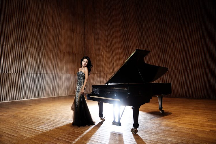 Joyce Yang poses beside a grand piano in an elegant concert setting, marking her performance in the Omaha Symphony’s season finale. She wears a formal outfit, exuding confidence and grace.