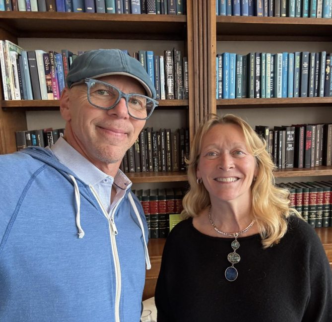 Jeff Koterba and Denise Natali smile for selfie in front of a bookcase. 