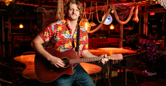 Actors perform a lively beachside scene from Escape to Margaritaville, dressed in tropical outfits with colorful lights and palm tree backdrops.