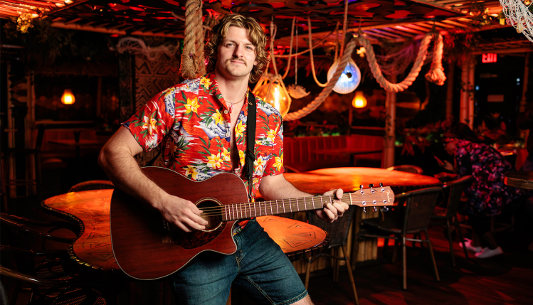Actors perform a lively beachside scene from Escape to Margaritaville, dressed in tropical outfits with colorful lights and palm tree backdrops.