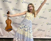 Classical Kid, Sara, poses in front of an Omaha Conservatory of Music Backdrop. She has a huge smile and holds her violin and has her other hand in the air.