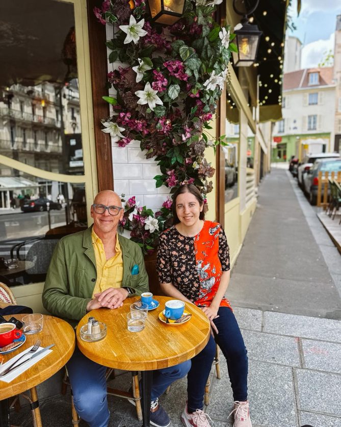 Jeff and Alicia sit at a table outside of a cafe in France. 