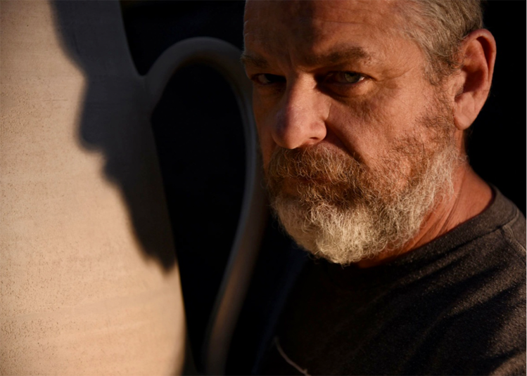 Rodney Tague smiling while posing in front of his pottery work, representing Clay Contours.