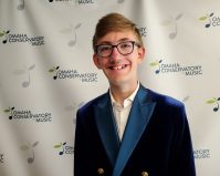 Andy Brink, the KVNO Classical Kid for September, stands smiling in front of Omaha Conservatory of Music banner.