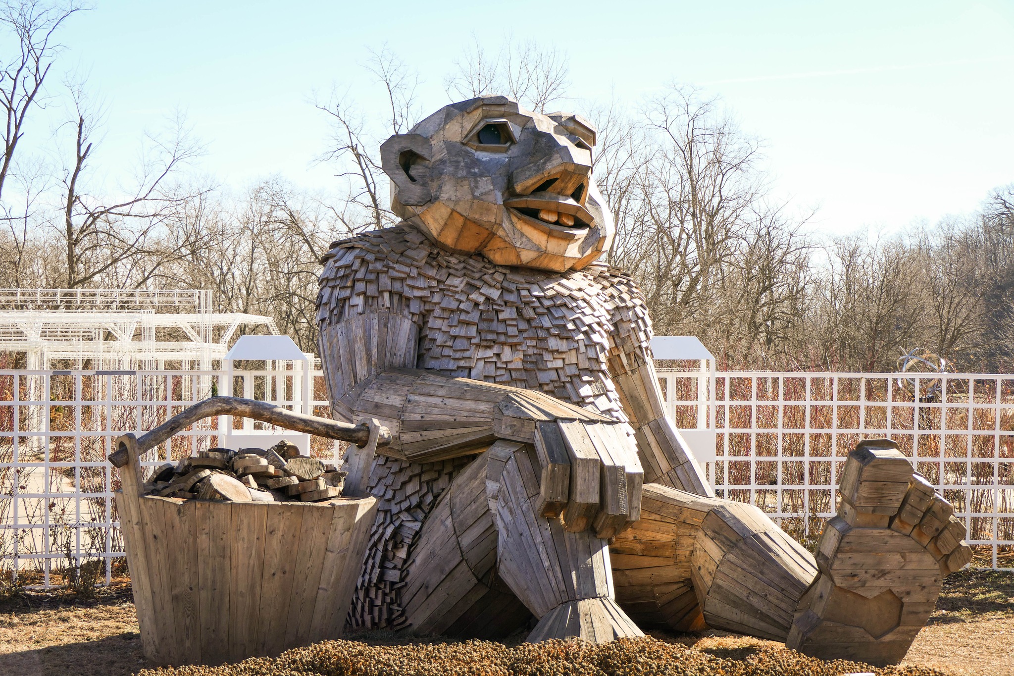 Large wooden troll sculpture made from reclaimed wood stands along a garden path, towering above visitors and greenery, part of Thomas Dambo’s TROLLS: Save the Humans outdoor exhibition.