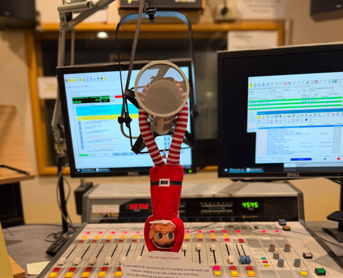 Photo of an Elf on the Shelf stuffed doll hanging from the microphone of the main studio at KVNO. 