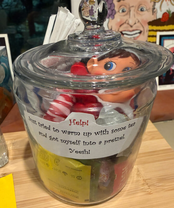 Elf on the Shelf stuffed elf tied up in a pretzel in a cookie jar filled with tea bags and snacks. 