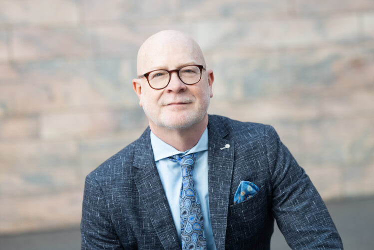 Portrait of Jack Becker, executive director of the Joslyn Art Museum, posing in a blue suit with a happy expression.