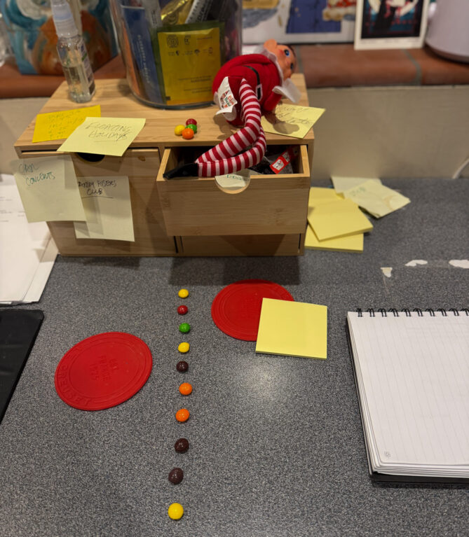 Elf on the Shelf--called Ivanhoe at KVNO--is seen in an open drawer of a small cabinet with with papers and post-it notes scattered about. Also, ,there's a trail of Skittles on the desk leading to the Elf.