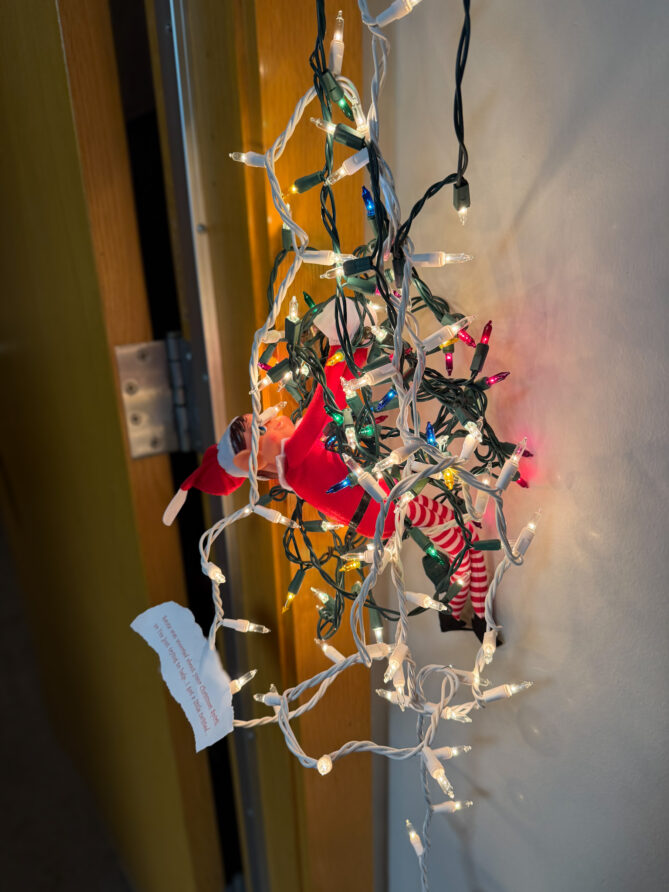 Elf on the Shelf named Ivanoe at KVNO all tangled up in Christmas lights outside of the door to the studio. A message on the elf says: "Santa was worried about your Christmas Spirit...so I'm just trying to help...I got a little twisted..."