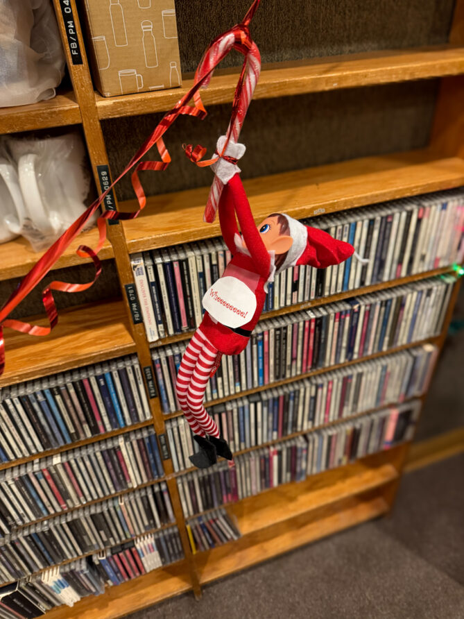 Ivanhoe, the elf on the shelf at KVNO is seen holding a candy cane which is resting on a long thread of red ribbon as though he is zip-lining.