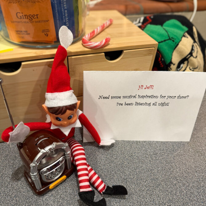 Elf on the shelf holding onto the antenna of an old-fashioned looking radio with a note that say: Hi Jeff! Need some musical inspiration for your show? I've been listening all night!