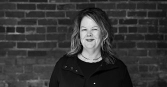 Maggie Wood, Executive Director of Film Streams, stands in front of a brick wall with a small smile in a black and white portrait.