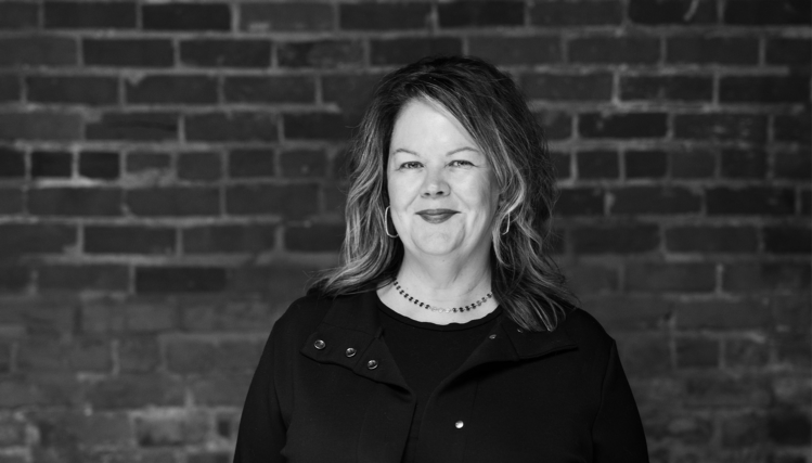 Maggie Wood, Executive Director of Film Streams, stands in front of a brick wall with a small smile in a black and white portrait.