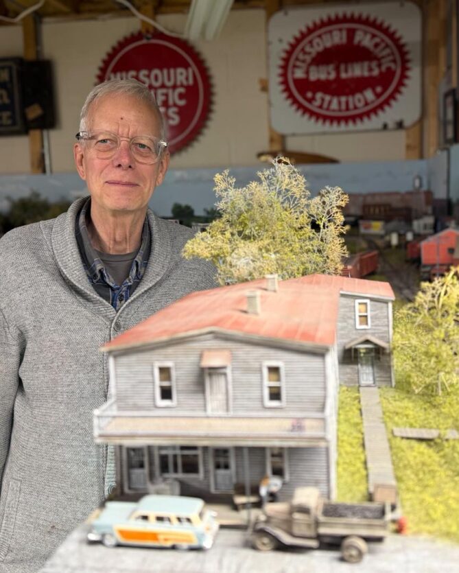 Charlie Duckworth stands next to a model building with model cars in front of it. 