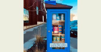 BLUEBARN Theatre's Little Free Pantry, painted blue with a clear glass door and a 3D printed sign that spells out BLUEBARN on the top, is shown photographed outside the theatre with a cream border.