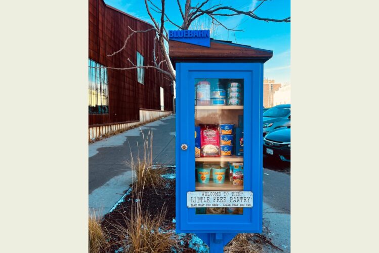 BLUEBARN Theatre's Little Free Pantry, painted blue with a clear glass door and a 3D printed sign that spells out BLUEBARN on the top, is shown photographed outside the theatre with a cream border.