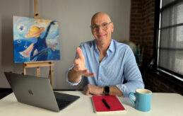 Jeff Koterba motioning toward you, the viewer, sitting at a desk with an open laptop, notebook and pen, and a coffee cup. In the background is a painting on an easel.