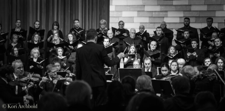 Black-and-white photograph taken from behind Maestro Barron Breland as he conducts an orchestra during a performance of Beethoven-77, his arms raised mid-gesture before the ensemble.
