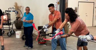 Three artists at Hot Shops Art Center cut and shape pieces of melted glass, carefully reassembling them into the form of an inauguration ribbon inside a working studio.