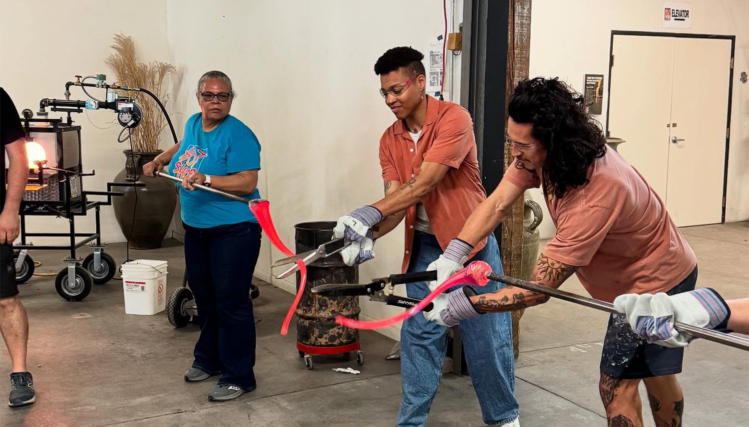 Three artists at Hot Shops Art Center cut and shape pieces of melted glass, carefully reassembling them into the form of an inauguration ribbon inside a working studio.