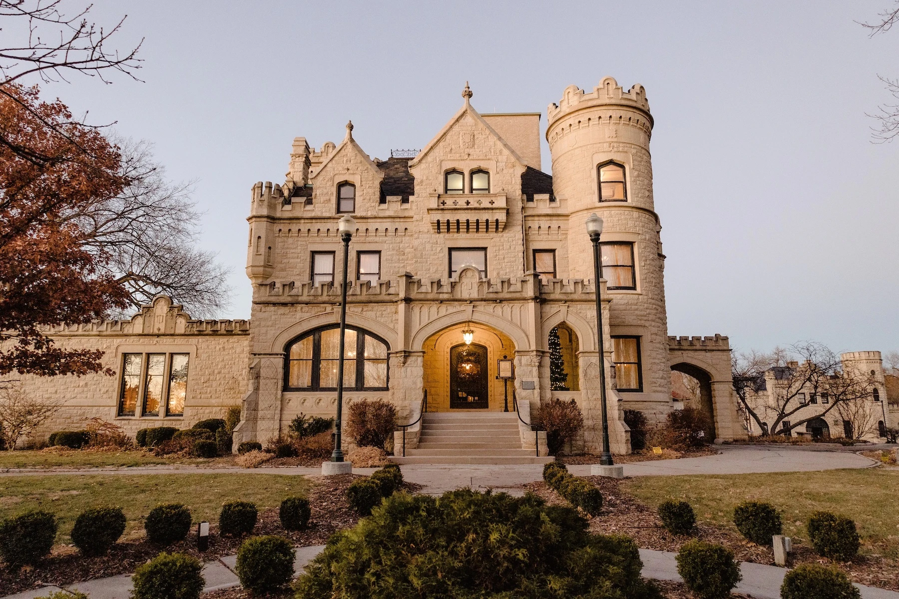 Photo of Joslyn Castle & Gardens in Omaha at sunset, the historic stone mansion warmly illuminated by golden light while the sky glows with soft orange and pink tones above the surrounding gardens.