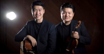 A professionally photographed portrait of ArcoStrum members TY Zhang and Strauss Shi standing against a dark background while holding a guitar and a violin.