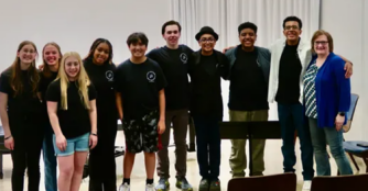 A diverse group of ten participants from Music Marathon for Music Education pose together inside a recital hall, holding instruments and smiling after a performance.