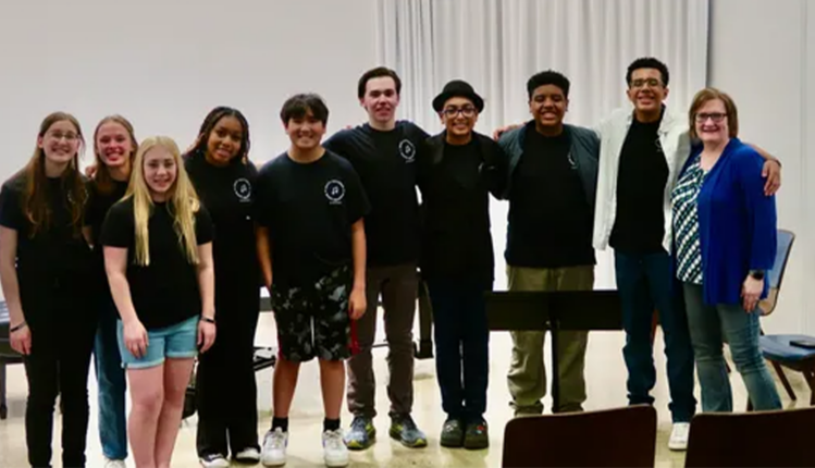 A diverse group of ten participants from Music Marathon for Music Education pose together inside a recital hall, holding instruments and smiling after a performance.