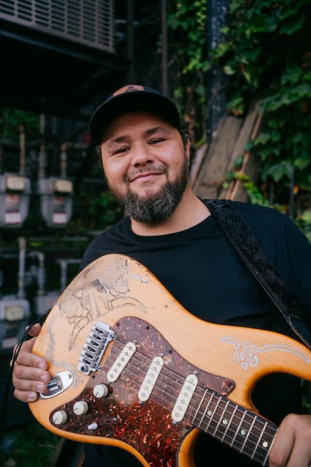 Portrait of Omaha blues musician Hector Anchondo standing with his guitar. Anchondo faces the camera with a relaxed expression while holding his instrument, reflecting his career as a guitarist, singer and songwriter.