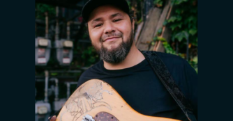 Hector Anchondo poses with his guitar and a light smile.